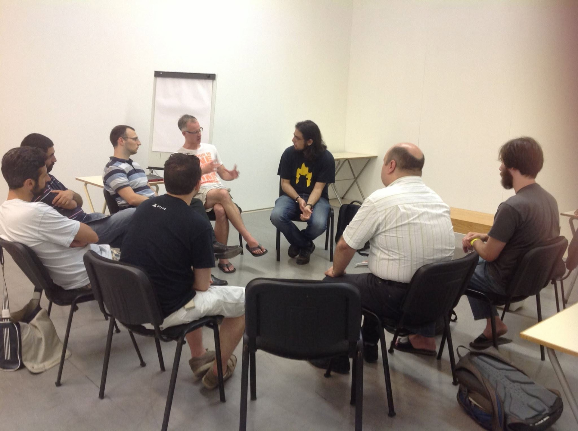 A group of people seated in a circle of chairs, in discussion in a white room
